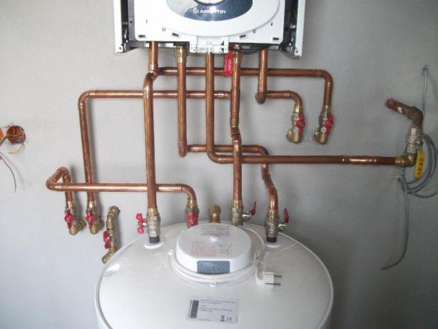 Biały zasobnik ciepłej wody z podłączonymi miedzianymi rurami i zaworami, zamontowany pod piecem grzewczym na szarej ścianie.