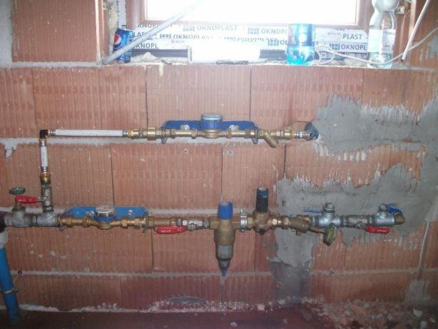 Surowe ściany z cegły z zamontowanymi rurami wodnymi, zaworami i licznikiem, widoczne napoje w tle na parapecie okna.