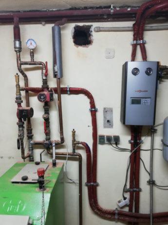 Piwnica z zainstalowanym kotłem grzewczym Viessmann, rurami miedzianymi i zielonym zbiornikiem, widoczne manometry i zabezpieczenia, ściany z widocznymi przewodami i puszkami elektrycznymi.