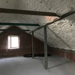 Poddasze w Krasnem po ociepleniu pianką, widoczne krokwie i ściany pokryte białą izolacją, na podłodze leżą czarne rury wentylacyjne.