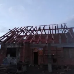 Budowa domu jednorodzinnego w trakcie wznoszenia więźby dachowej, widoczne krokwie i murłat, ściany z czerwonej cegły, pochmurne niebo w tle.