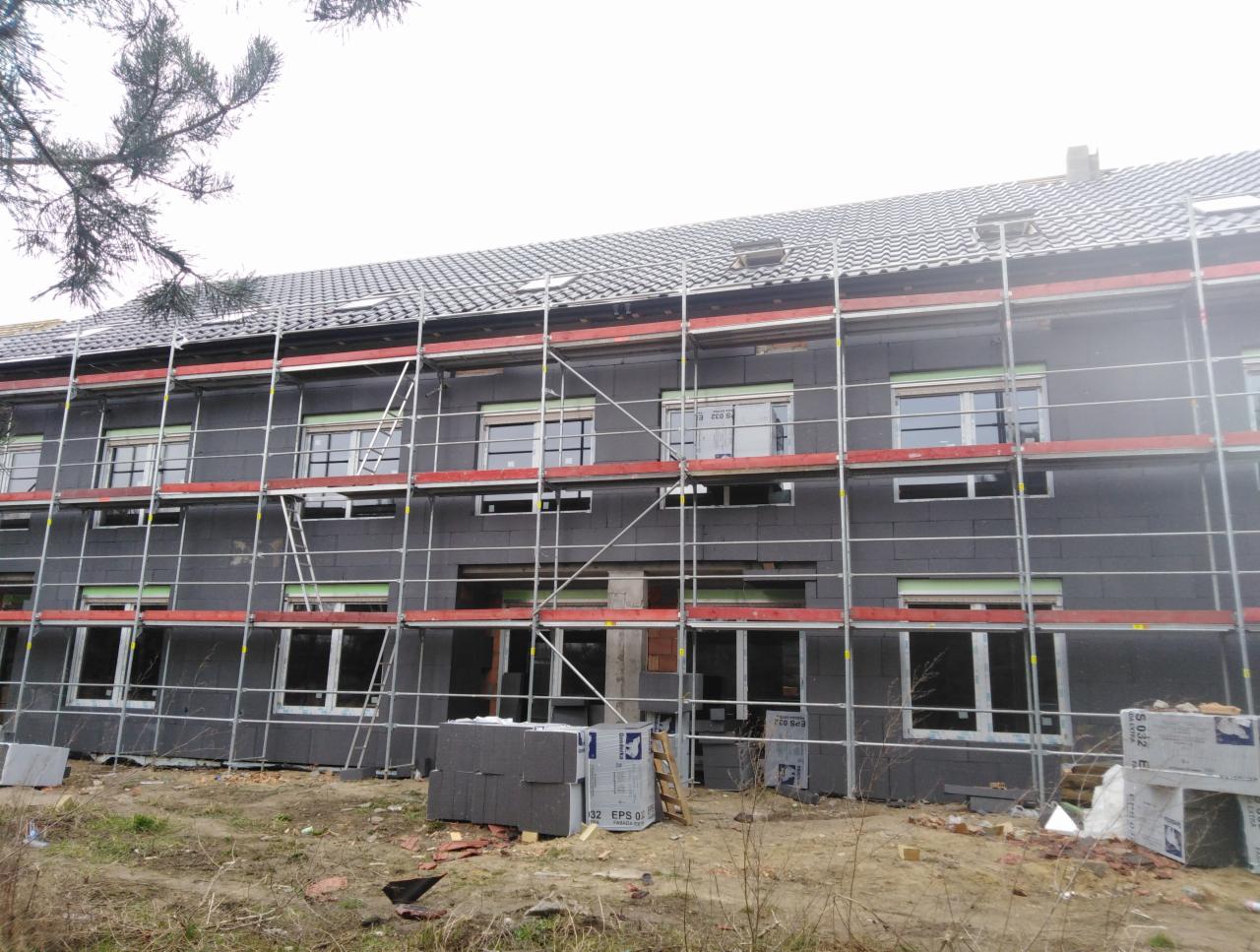 Budynek wielorodzinny w trakcie prac remontowych; widoczne rusztowania, ocieplenie ścian zewnętrznych styropianem grafitowym, dach pokryty szarą dachówką karpiówką, okna z częściowo zasłoniętymi...