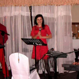 Trzyosobowy zespół muzyczny w czerwonych strojach, grający na keyboardach i śpiewający do mikrofonów na tle białych zasłon podczas występu.