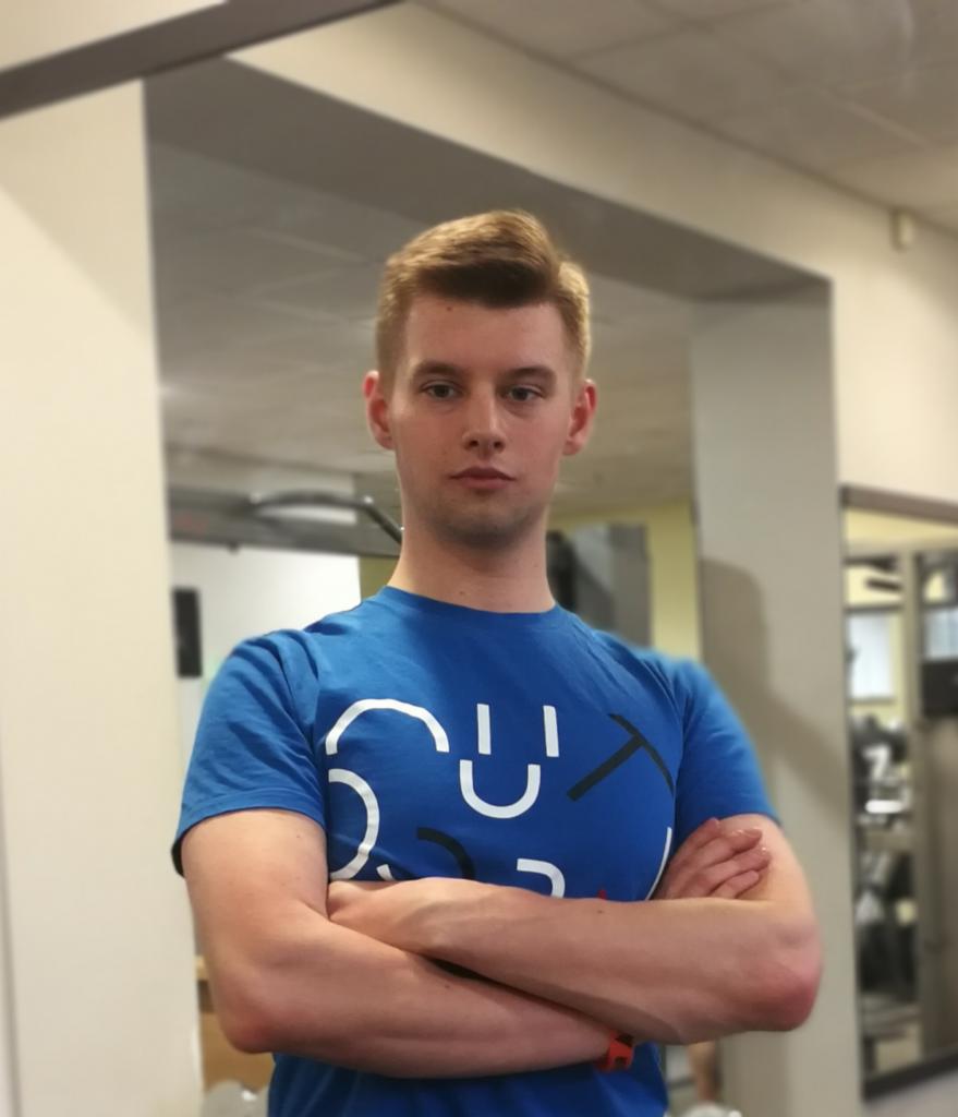 Młody mężczyzna z blond włosami w niebieskiej koszulce z nadrukiem, stoi z założonymi rękami na tle siłowni.