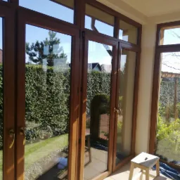 Przeszklony taras z drewnianą ramą okienną, widok na zadbany ogród z żywopłotem i dom z czerwoną dachówką, mały drewniany stołek w rogu pomieszczenia.