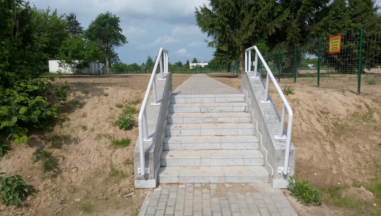 Betonowe schody z białymi barierkami prowadzące na górę, wykończone kostką brukową, otoczone ziemią i zielenią.