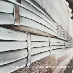 Szereg szarych przewodów elektrycznych ułożonych równolegle i przymocowanych metalowymi uchwytami do surowej, szarej ściany. Widoczne napisy na przewodach. W tle fragment konstrukcji sufitu.