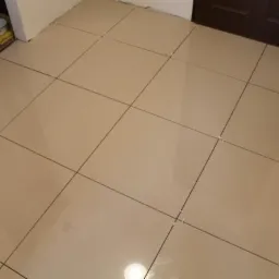 Beżowe, błyszczące płytki podłogowe ułożone w kwadratową siatkę, z widocznymi krzyżykami dystansowymi w spoinach, przy ścianie i brązowych drzwiach.