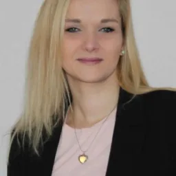 Portret blondynki w czarnej marynarce i różowej koszulce, z naszyjnikiem w kształcie serca, uśmiechającej się do kamery.