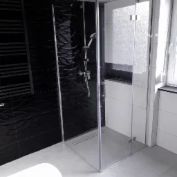 Nowoczesna kabina prysznicowa z czarnymi płytkami o falistej strukturze i grzejnikiem drabinkowym, kontrastująca z białymi płytkami i oknem.