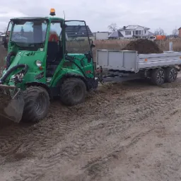 Zielona miniładowarka Avant 750 z otwartą kabiną i przyczepą wypełnioną ziemią na placu budowy, w tle nowe domy jednorodzinne.