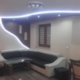 Pok&oacute;j dzienny z narożną sofą, szklanym stolikiem i oświetleniem LED wzdłuż ściany, uzupełniony o dekoracyjną lampę sufitową i minimalistyczne wnęki ścienne.