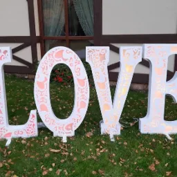 Duże, białe, podświetlane litery LOVE z ażurowym wzorem stojące na trawie przed budynkiem z oknami.