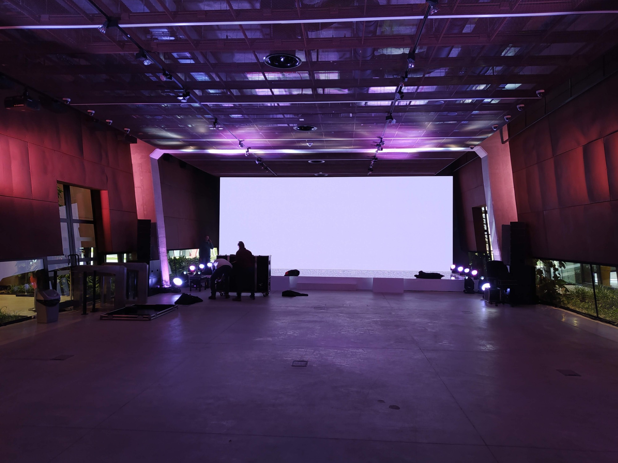 Przygotowania do wydarzenia w hali: montaż ekranu, oświetlenia i nagłośnienia przez ekipę techniczną w fioletowym oświetleniu.