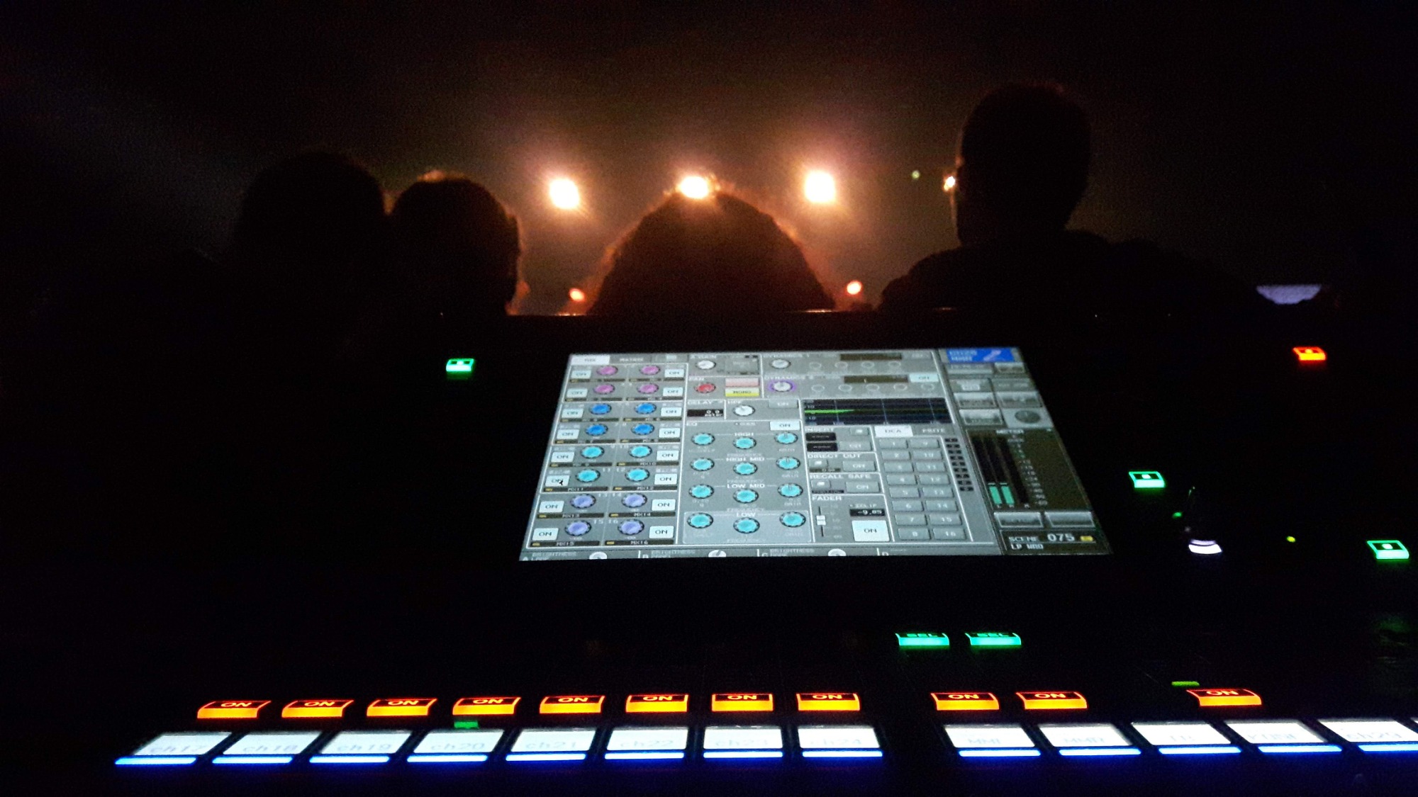 Konsola mikserska oświetlona diodami, w tle sylwetki osób i sceniczne światła, widok z perspektywy operatora.