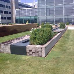 Zachara Lębryk - Nowoczesny taras na dachu budynku biurowego z drewnianymi elementami, zielonym trawnikiem i roślinami ozdobnymi, w tle widoczny pracownik w kamizelce odblaskowej.