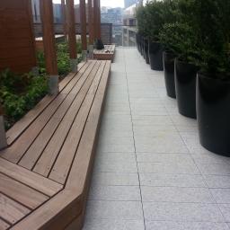 Zachara Lębryk - Drewniany taras na dachu budynku z roślinami w donicach i widokiem na miasto w tle.