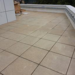 Zachara Lębryk - Wykończony taras na dachu budynku, wyłożony kwadratowymi płytami chodnikowymi w beżowym odcieniu, z widoczną metalową barierką przymocowaną do białego murku i fragmentem elewacji z oknem po lewej...