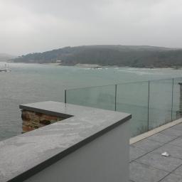 Zachara Lębryk - Nowoczesny taras z szarą, kwadratową nawierzchnią, przeszkloną balustradą i widokiem na morze w pochmurny dzień.