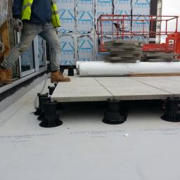 Zachara Lębryk - Montaż podniesionego tarasu na dachu: pracownik w butach roboczych wchodzi na taras z płyt betonowych, wspartych na regulowanych wspornikach, widoczne materiały budowlane i podnośnik koszowy w tle.