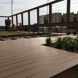 Zachara Lębryk - Montaż tarasu kompozytowego na dachu budynku, widoczna konstrukcja drewniana pergoli, trawa w rolkach i pracownik w kamizelce odblaskowej nadzorujący prace na tle miejskiego krajobrazu.