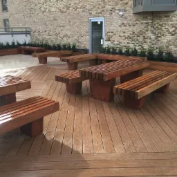 Umeblowany taras na dachu budynku z drewnianymi ławkami i stołami, otoczony zielenią w donicach, z widoczną elewacją z cegły w tle.