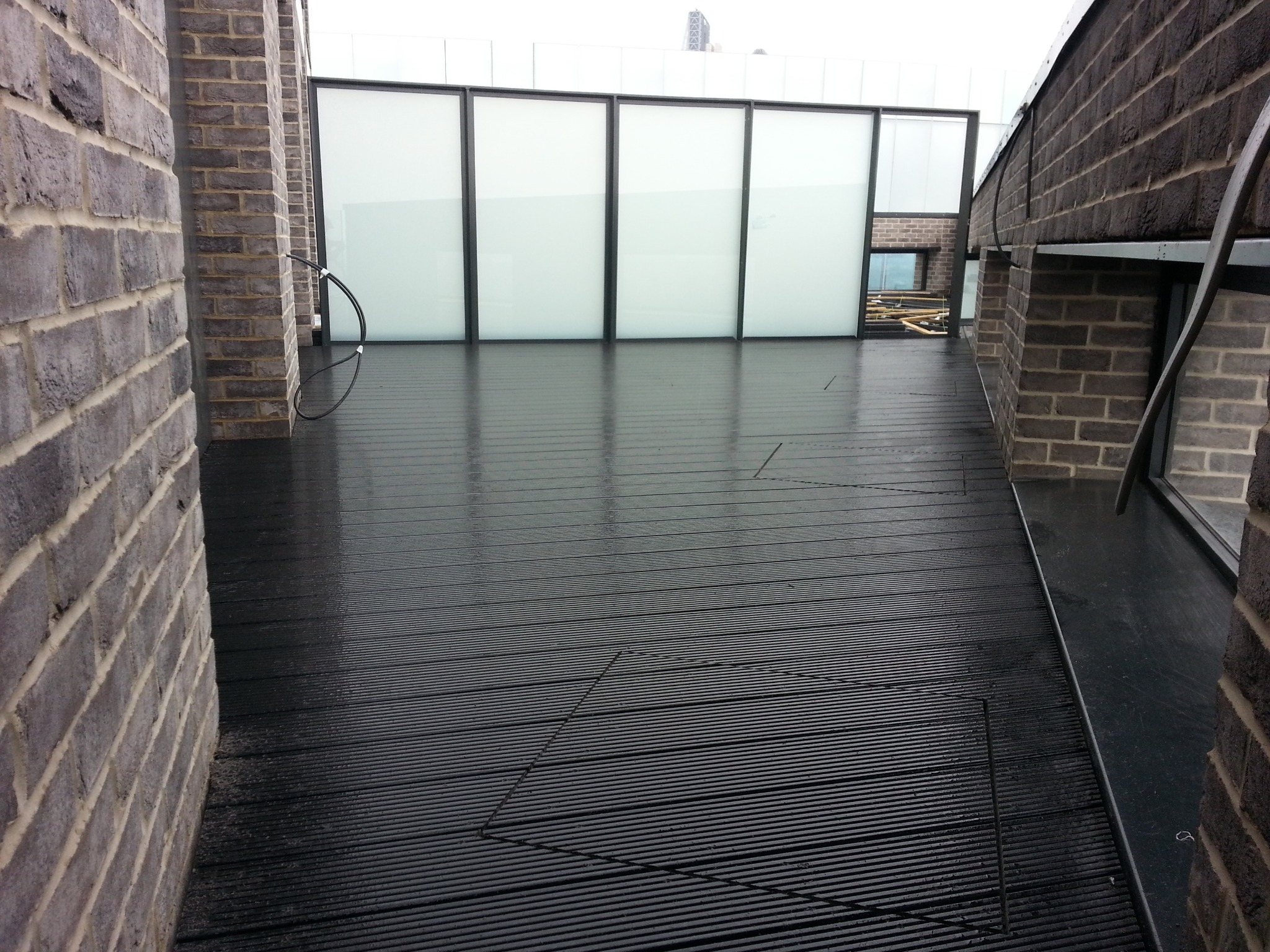 Czarny taras z desek kompozytowych na dachu budynku, otoczony ceglanymi ścianami i przeszklonymi panelami, widok z niskiej perspektywy, po deszczu.