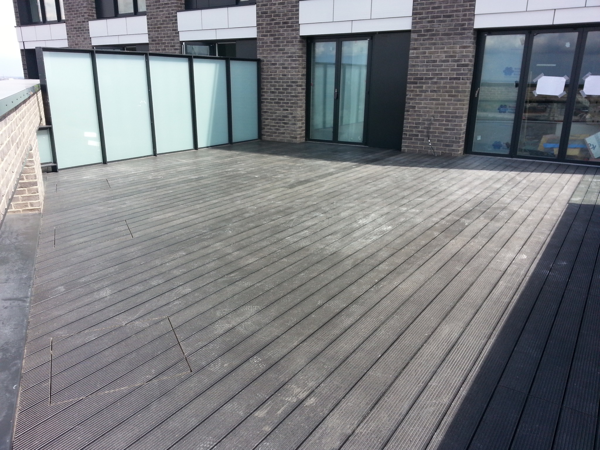 Szary taras z desek kompozytowych na dachu budynku, z widoczną szklaną balustradą i fragmentem elewacji z cegły.
