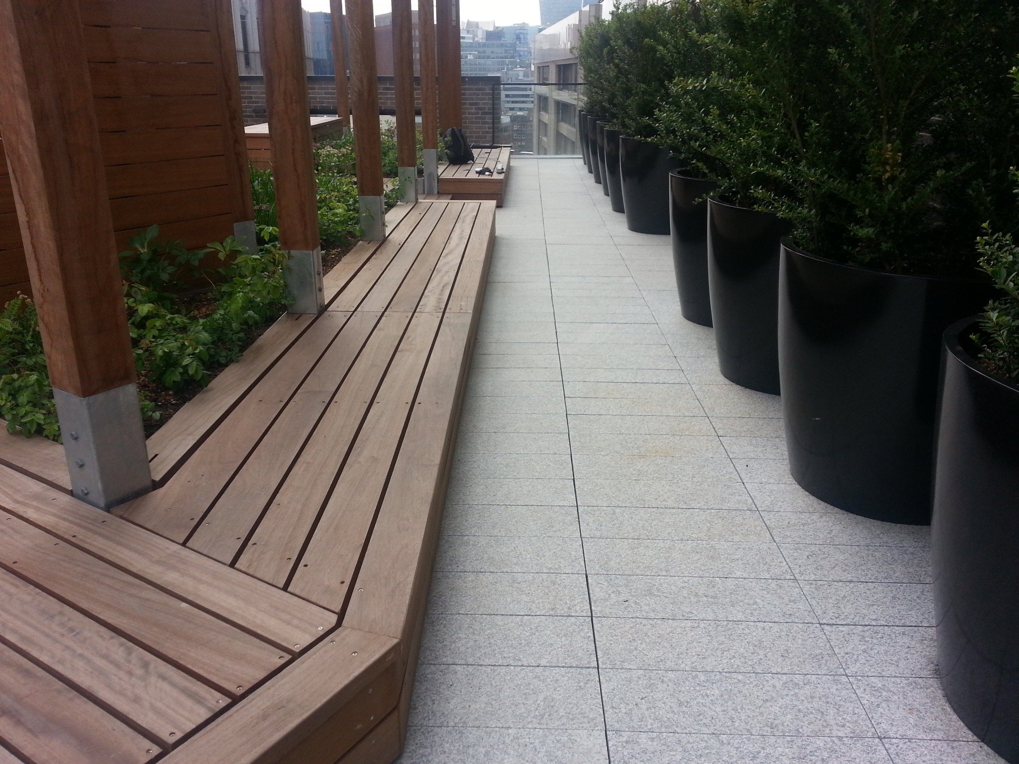 Drewniany taras na dachu budynku z roślinami w donicach i widokiem na miasto w tle.