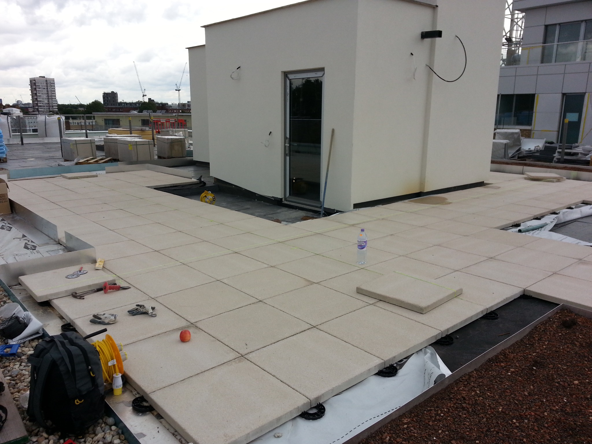 Montaż płyt tarasowych na dachu budynku, widoczne narzędzia, poziomica, rękawice robocze, plecak, butelka wody, niedokończona powierzchnia tarasu z widocznymi elementami konstrukcyjnymi i izolacją,...