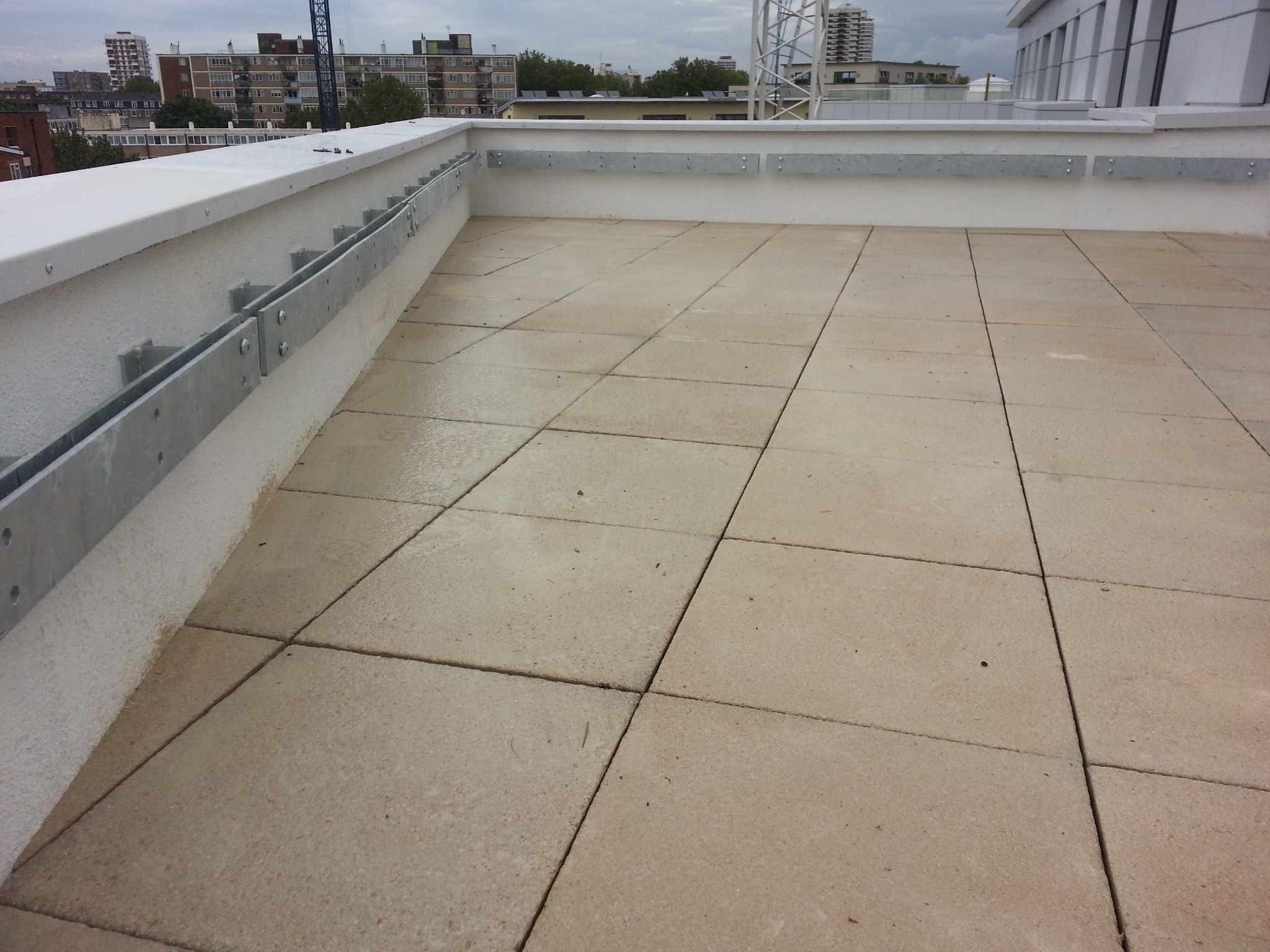 Płaski dach pokryty kwadratowymi płytami, widoczne metalowe wsporniki przymocowane do ściany attyki, w tle zabudowa miejska i dźwig budowlany.