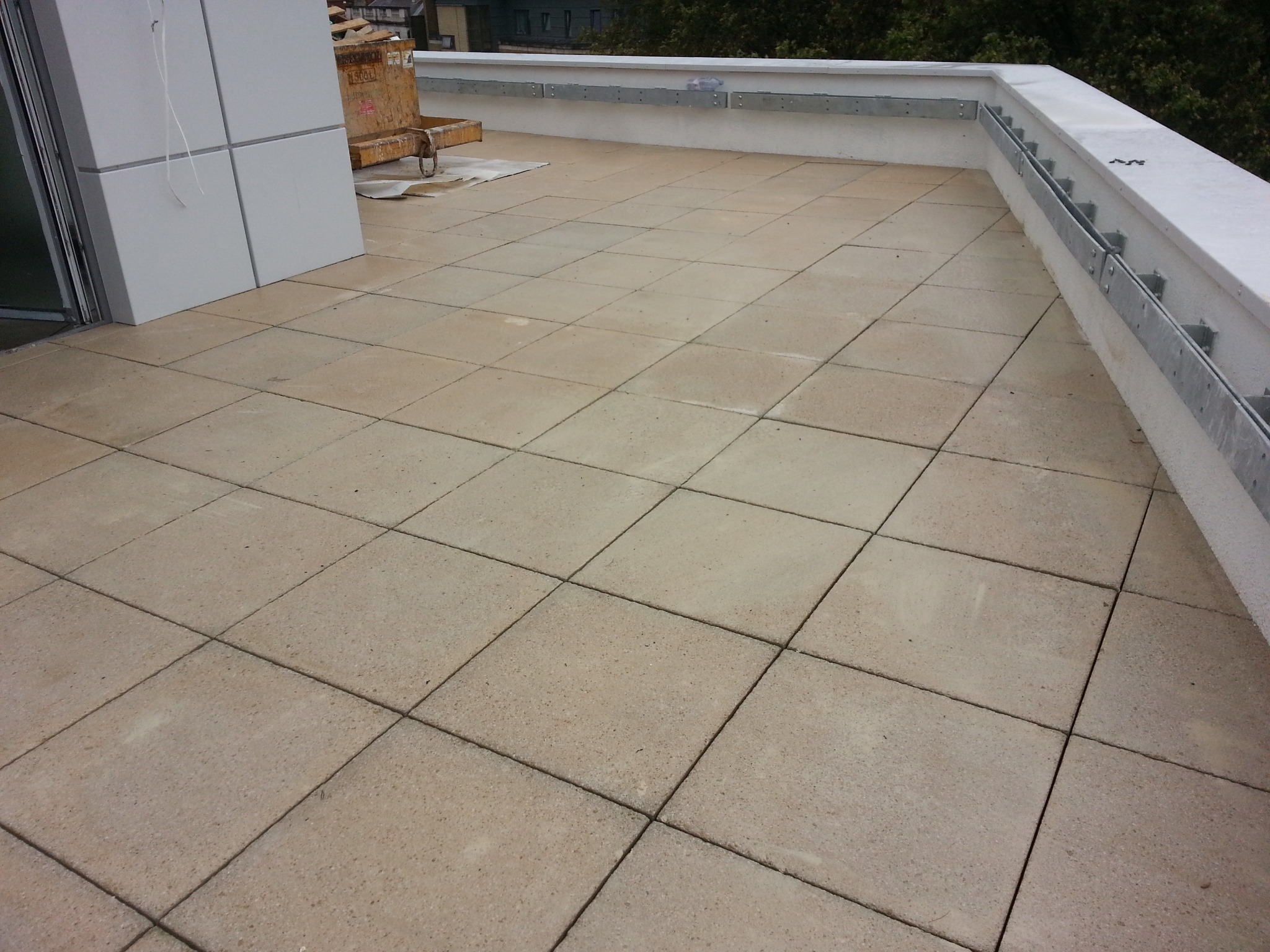 Wykończony taras na dachu budynku, wyłożony kwadratowymi płytami chodnikowymi w beżowym odcieniu, z widoczną metalową barierką przymocowaną do białego murku i fragmentem elewacji z oknem po lewej...