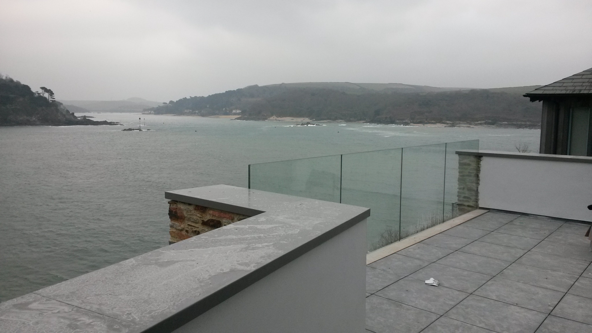 Nowoczesny taras z szarą, kwadratową nawierzchnią, przeszkloną balustradą i widokiem na morze w pochmurny dzień.