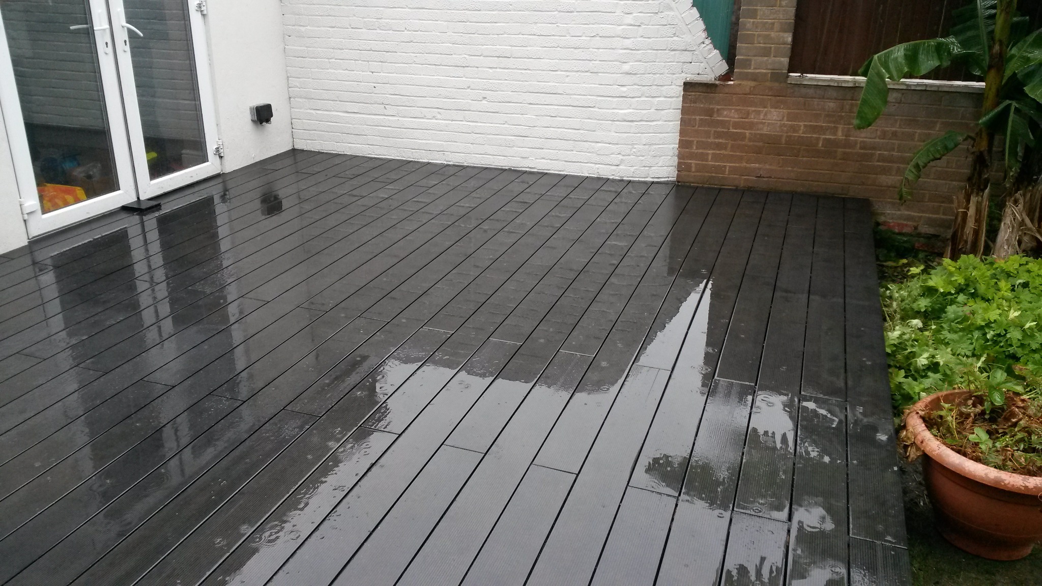 Szare deski tarasowe, mokre od deszczu, przylegające do białej ściany budynku z przeszklonymi drzwiami i ceglanego murku z zielenią.