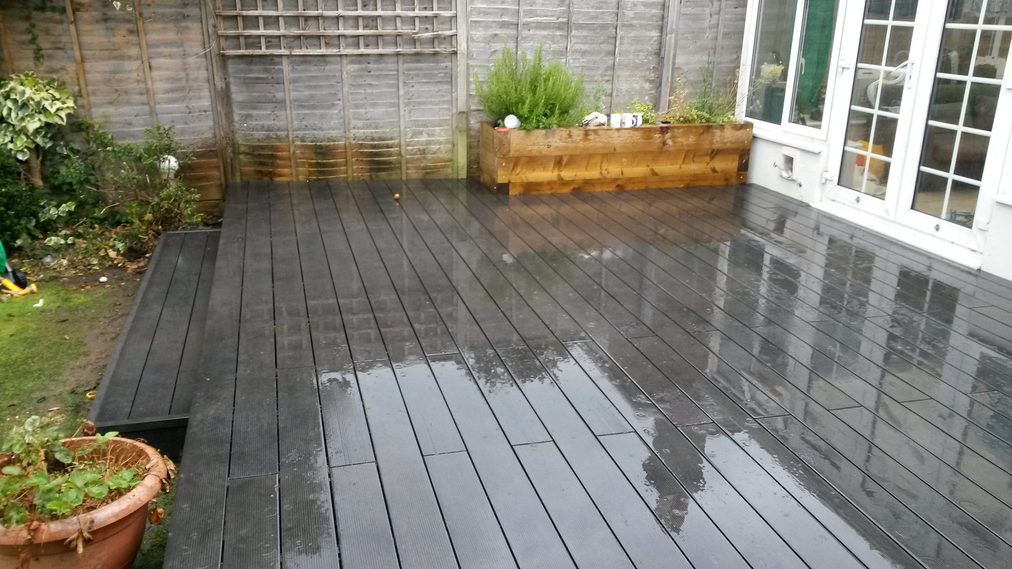 Szary taras kompozytowy po deszczu, z drewnianą skrzynią na rośliny i białymi drzwiami tarasowymi w tle.