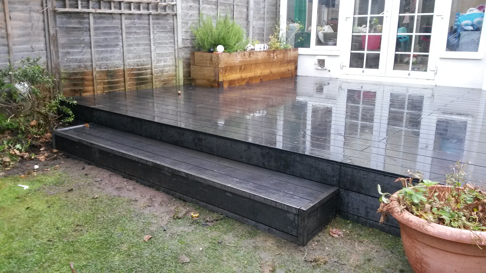 Czarny taras kompozytowy z dwoma stopniami, mokry od deszczu, z drewnianą skrzynią na rośliny i białymi drzwiami tarasowymi w tle.