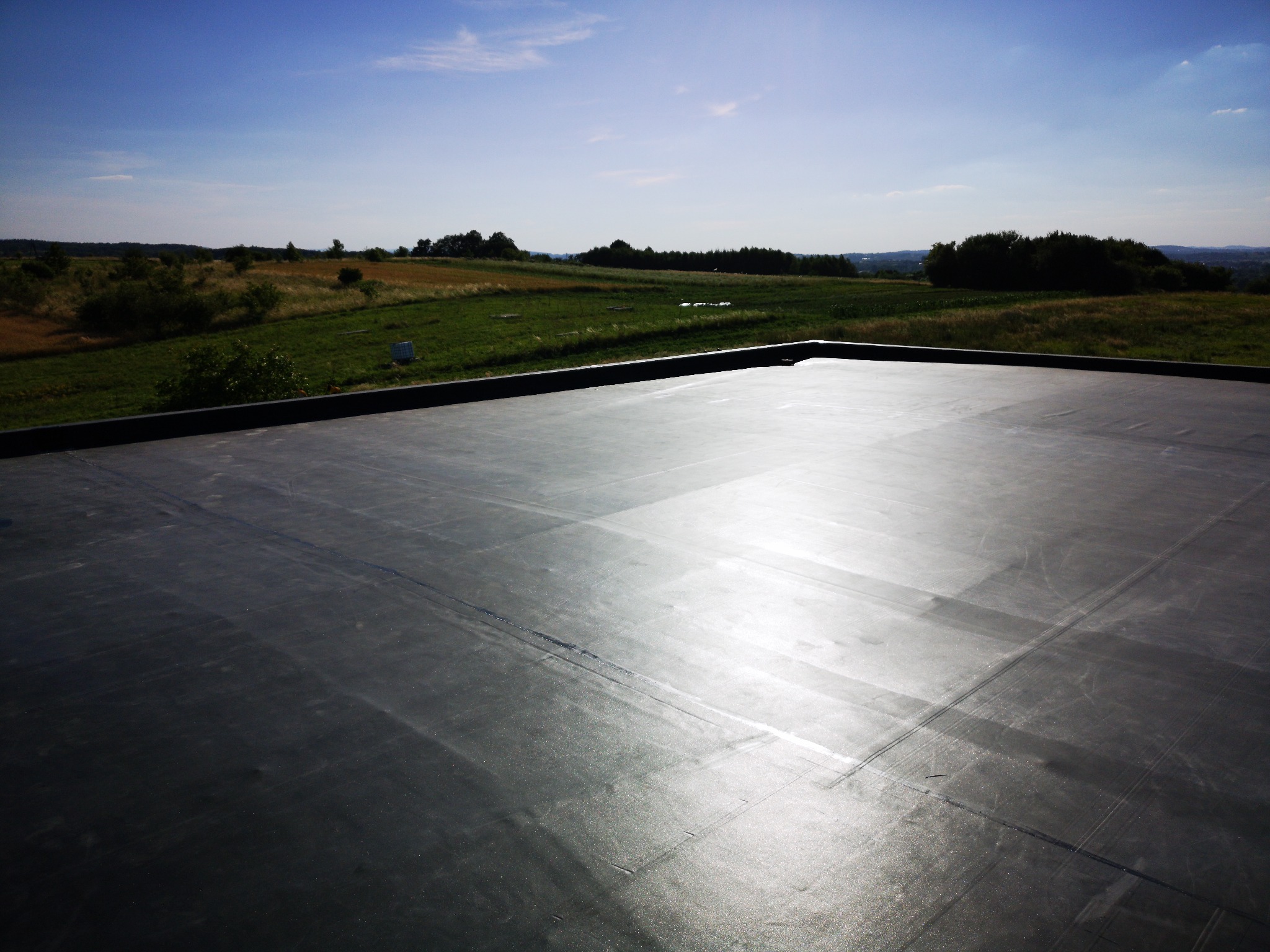 Czarna, płaska powierzchnia dachu z papy termozgrzewalnej z widokiem na zielone pola i lasy w oddali, w słoneczny dzień.