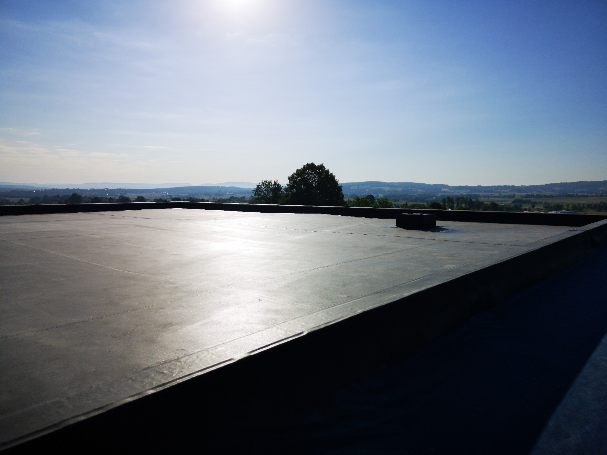 Płaski dach pokryty czarną membraną bitumiczną z widokiem na odległy krajobraz, błękitne niebo i słońce w górnym rogu.