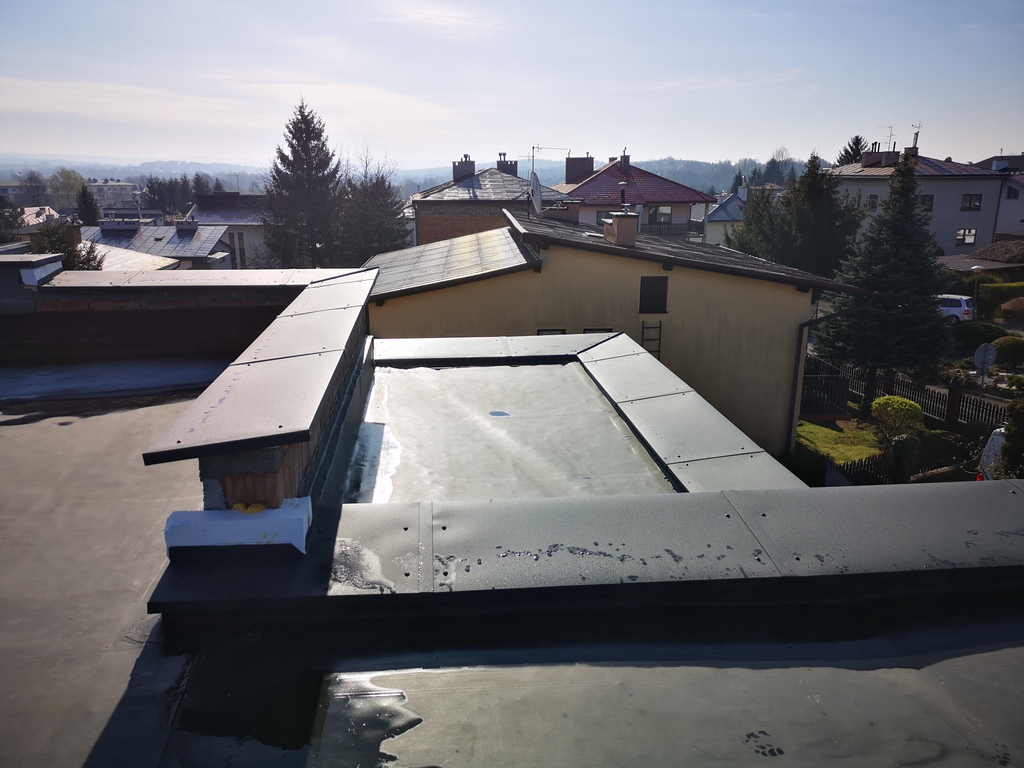 Widok z góry na płaski dach z obróbką blacharską, w tle zabudowa jednorodzinna i drzewa w słoneczny dzień.