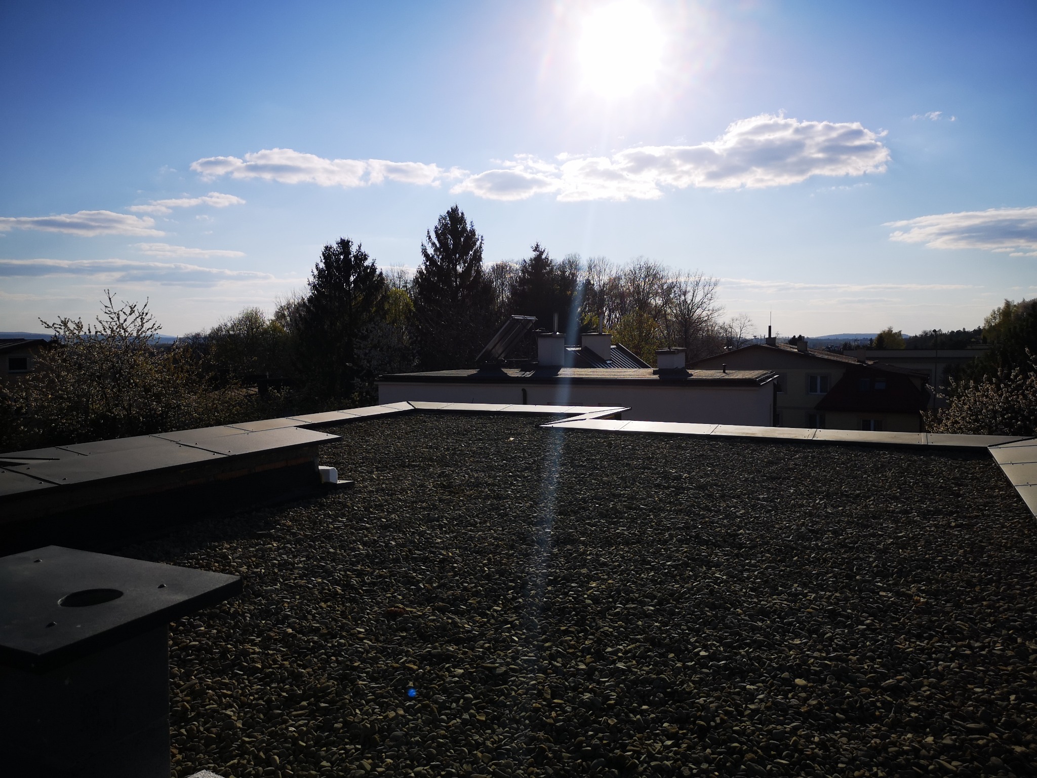 Dach pokryty drobnym żwirem, z widocznymi elementami konstrukcyjnymi na krawędziach, na tle błękitnego nieba z chmurami i jasnym słońcem, z widocznymi drzewami i budynkami w oddali.