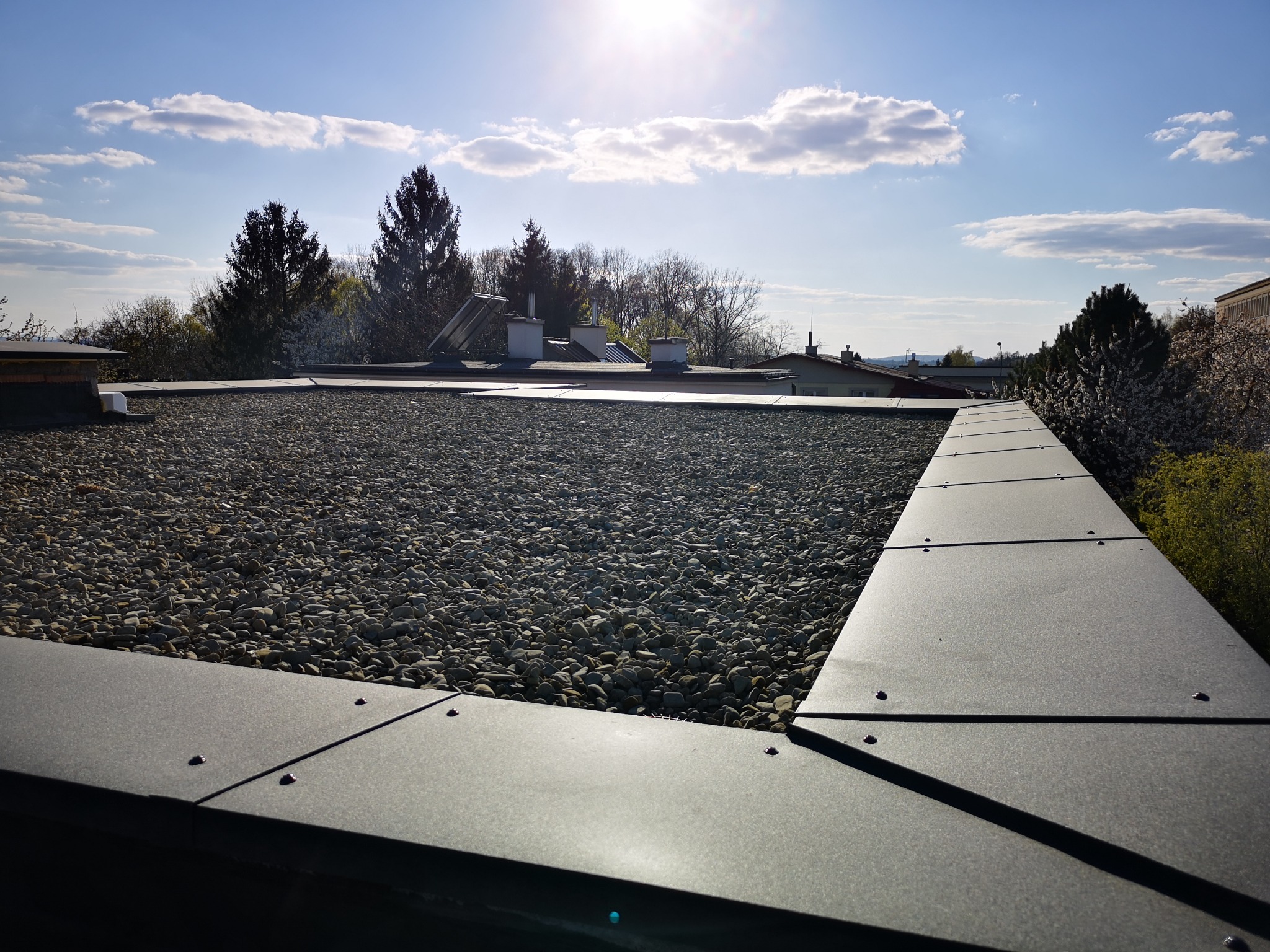 Płaski dach pokryty drobnymi kamykami, obramowany metalową listwą z widocznymi mocowaniami, w tle drzewa i błękitne niebo z chmurami.
