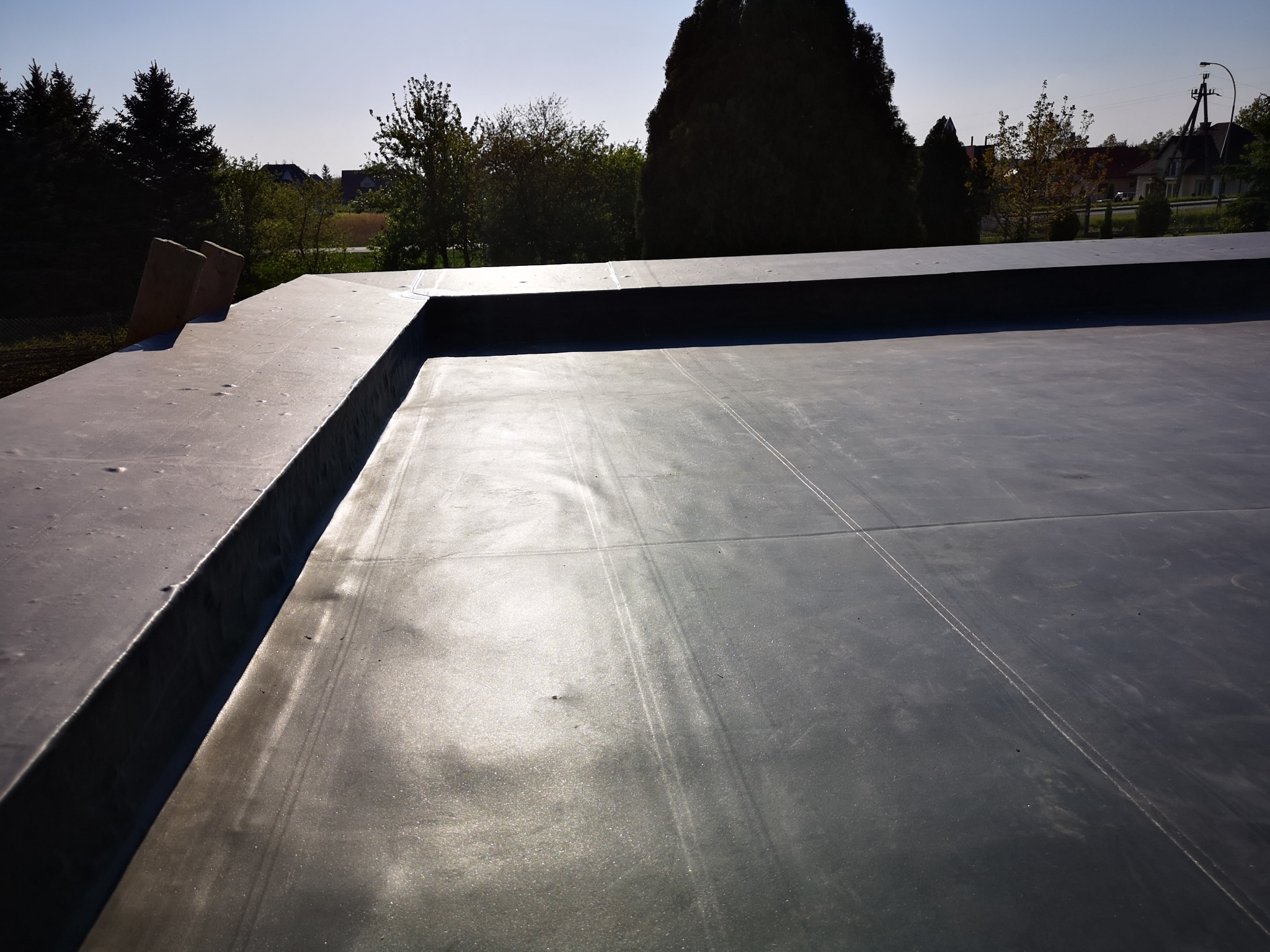 Widok z góry na płaski dach pokryty membraną, widoczne łączenia i obróbki blacharskie na krawędziach, w tle zieleń drzew i zabudowania.