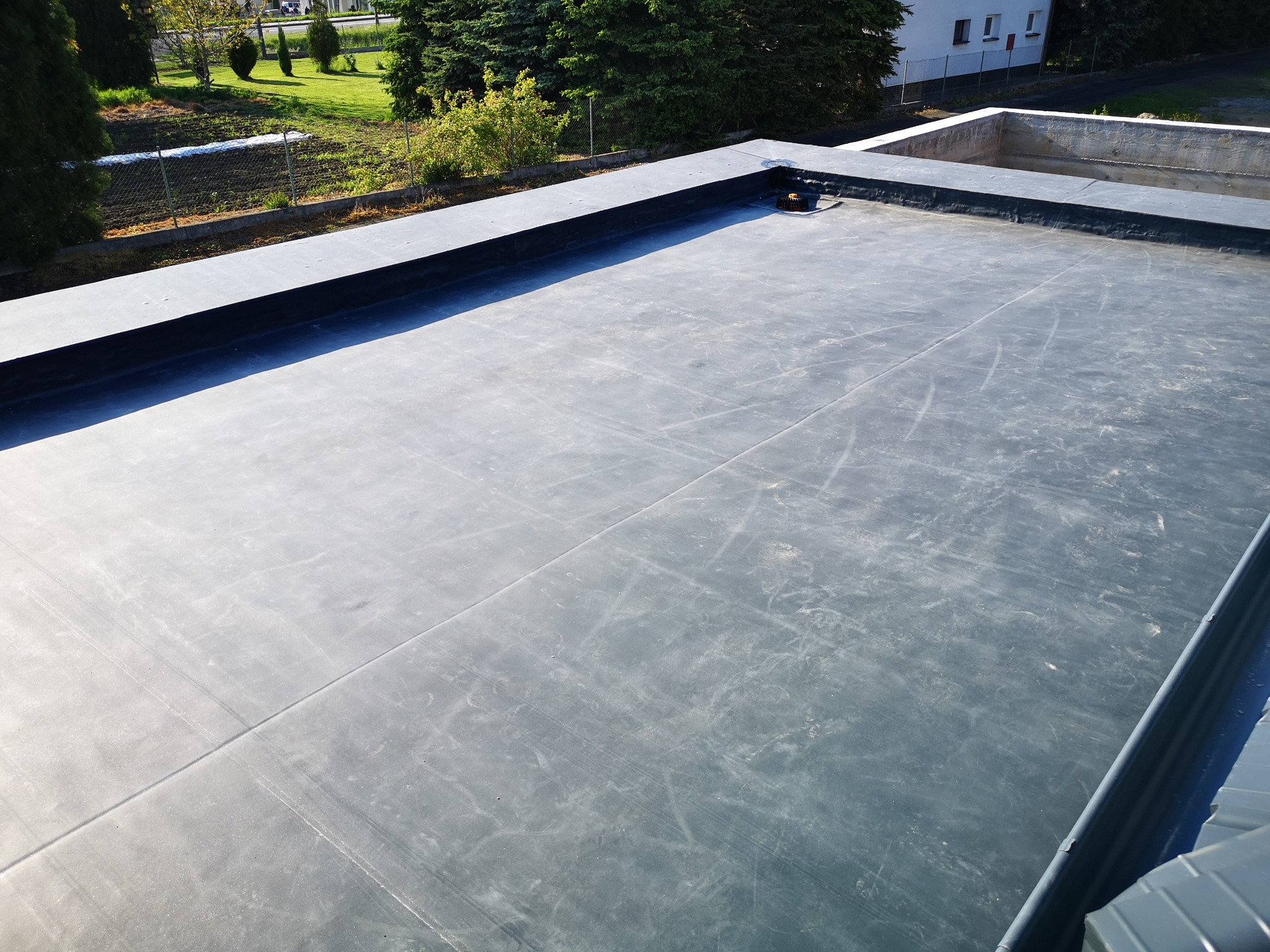 Płaski dach pokryty ciemną membraną bitumiczną, widoczne łączenia i detale wykończeniowe, w tle budynek mieszkalny i zieleń.
