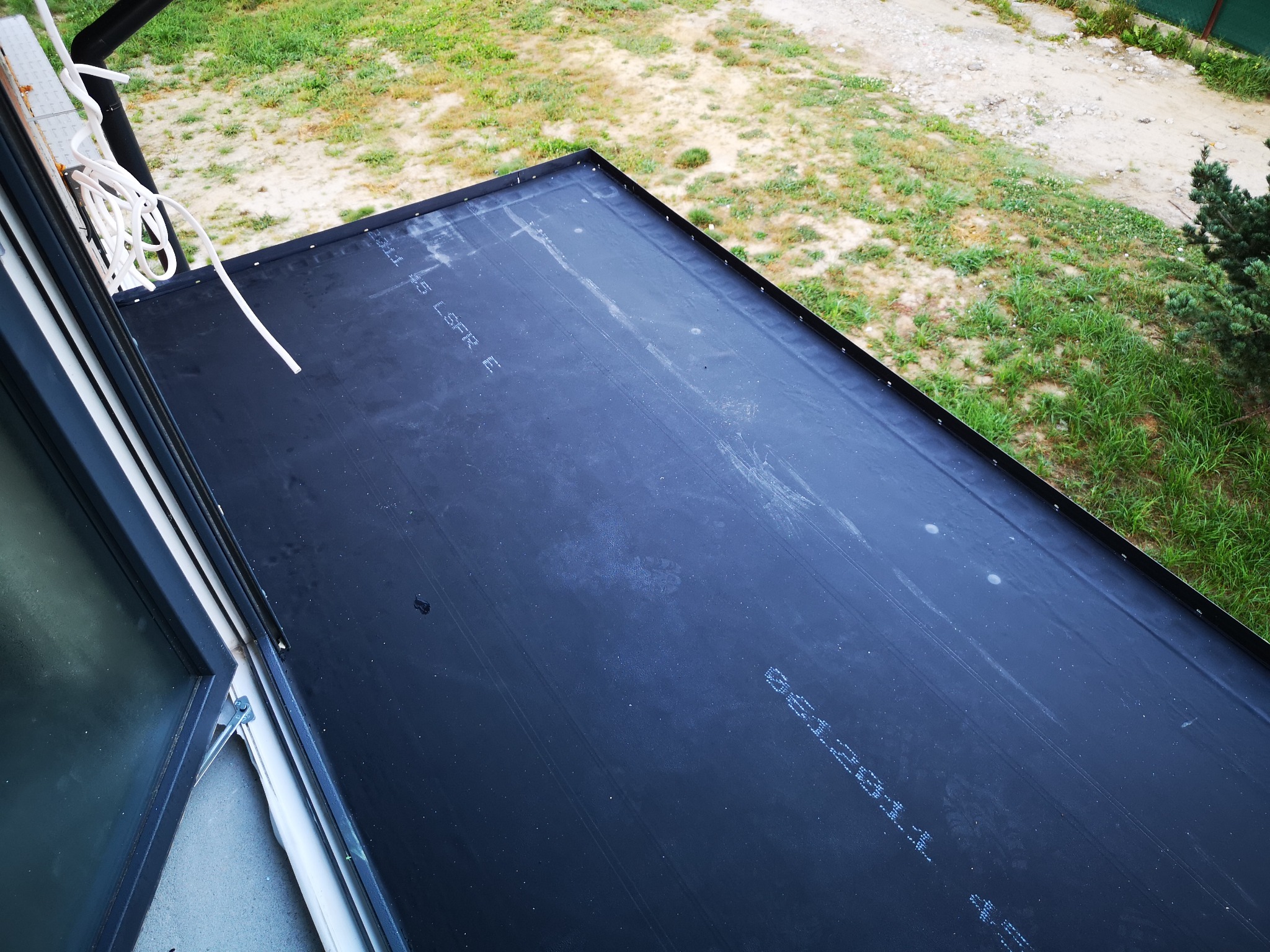 Widok z góry na fragment dachu pokrytego czarną membraną dachową z widocznymi napisami producenta, obok okno dachowe i fragment elewacji, w tle trawnik i drzewa.