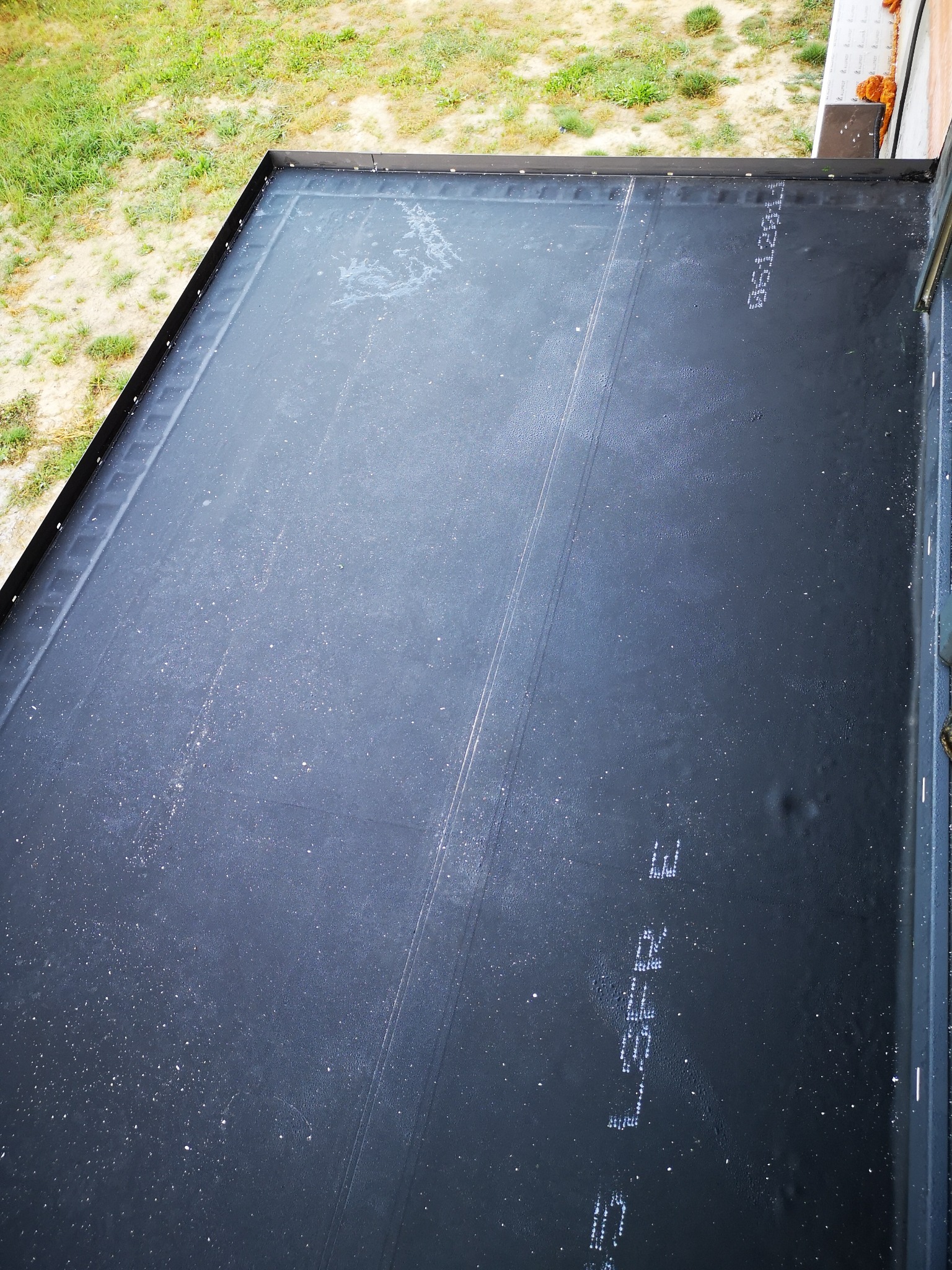 Widok z góry na fragment płaskiego dachu pokrytego czarną membraną, z widocznymi napisami producenta i metalową obróbką blacharską na krawędziach, obok fragmentu trawnika.