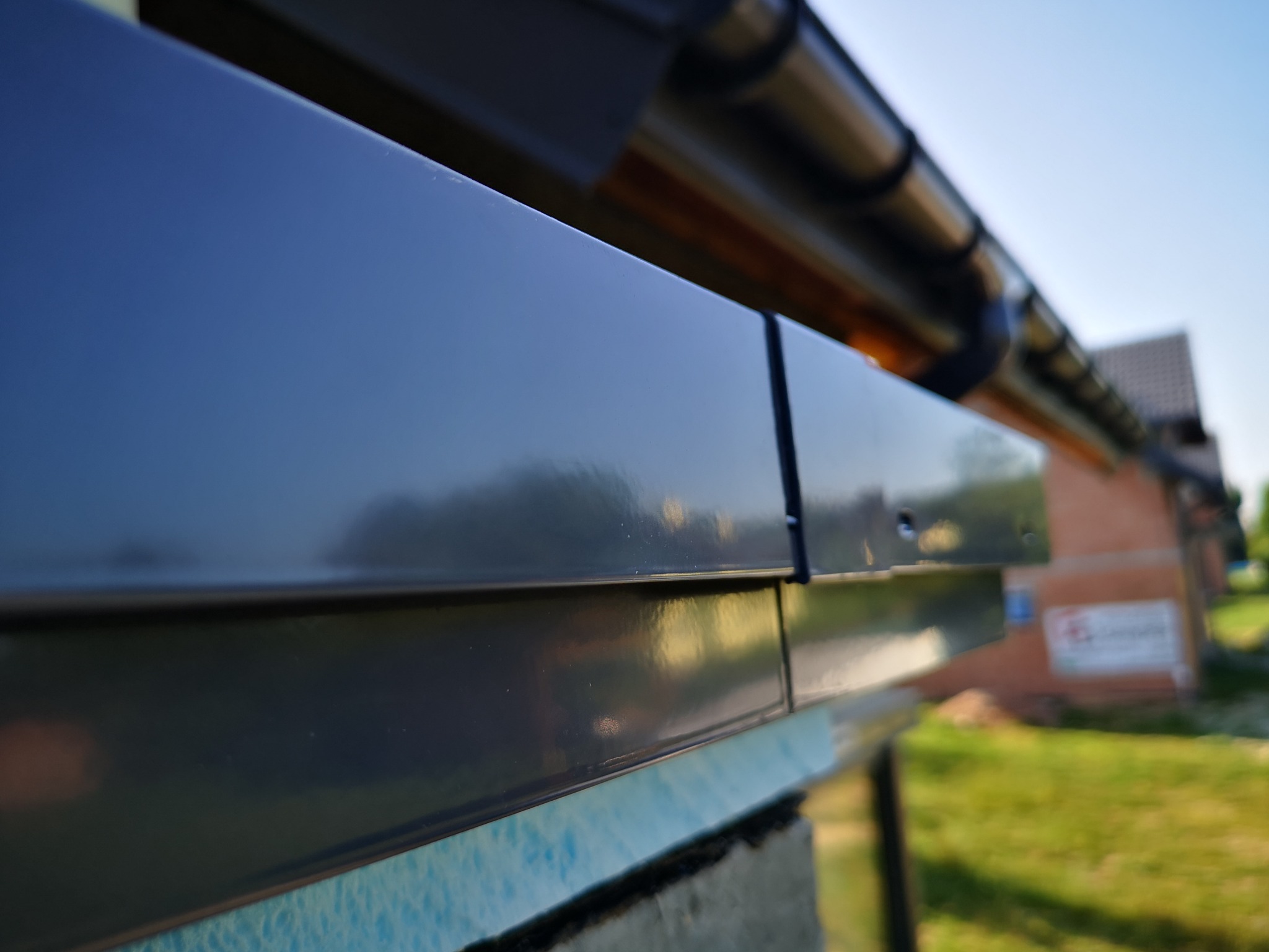 Detale nowej, błyszczącej, czarnej obróbki blacharskiej dachu, z widoczną rynną i fragmentem budynku w tle, wykonane z bliska.