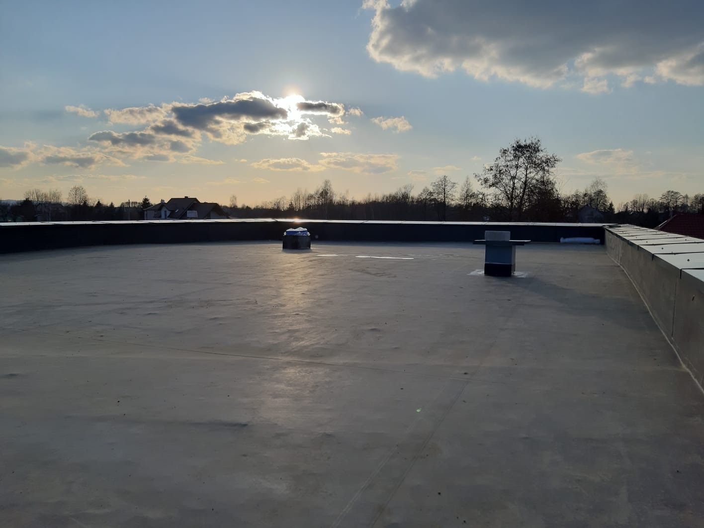 Płaski dach pokryty papą w Rzeszowie, widoczne kominki wentylacyjne, w tle zabudowa jednorodzinna i zachodzące słońce.