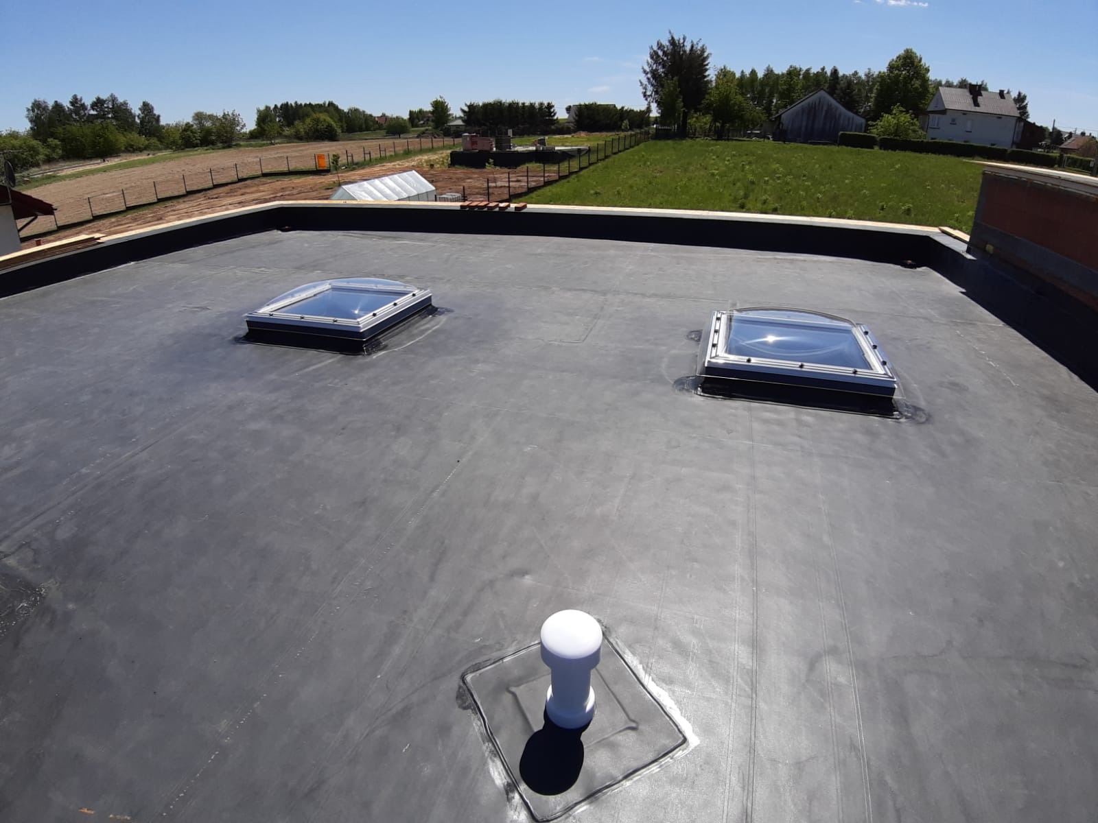 Płaski dach pokryty czarną membraną z dwoma świetlikami i odpowietrznikiem, widok z góry na tle zieleni i zabudowań.