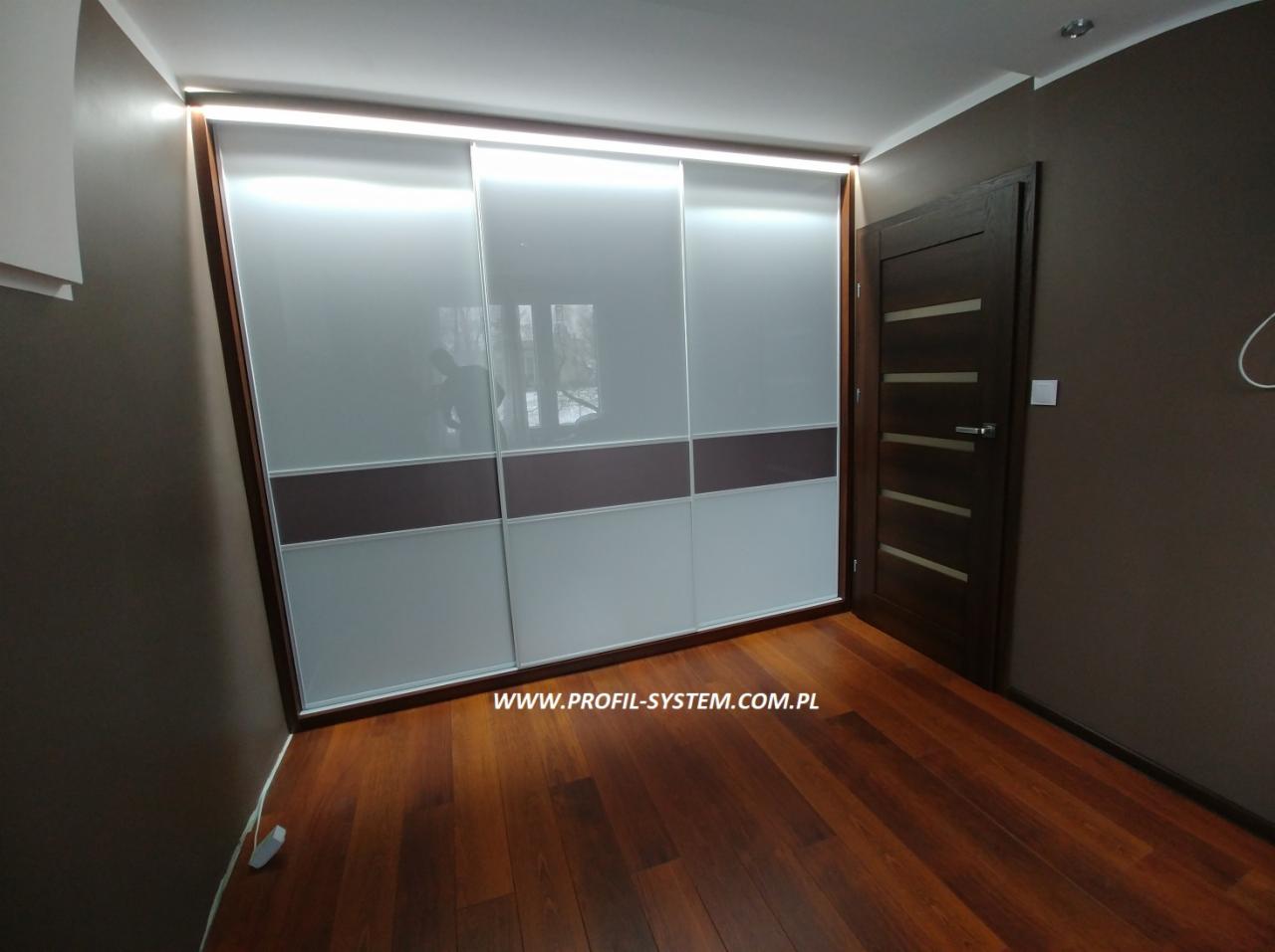 Przestronna szafa wnękowa z przesuwnymi, szklanymi drzwiami w odcieniach bieli i brązu, z oświetleniem LED u góry, zamontowana w pokoju w Katowicach z ciemnobrązowymi ścianami i drewnianą podłogą.