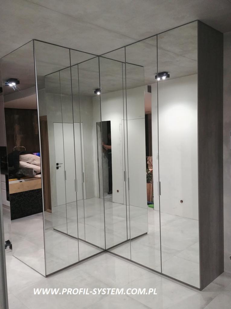 Nowoczesna szafa z lustrzanymi drzwiami, minimalistyczny design, widoczne odbicia wnętrza pokoju, szara ściana w tle, oświetlenie sufitowe.